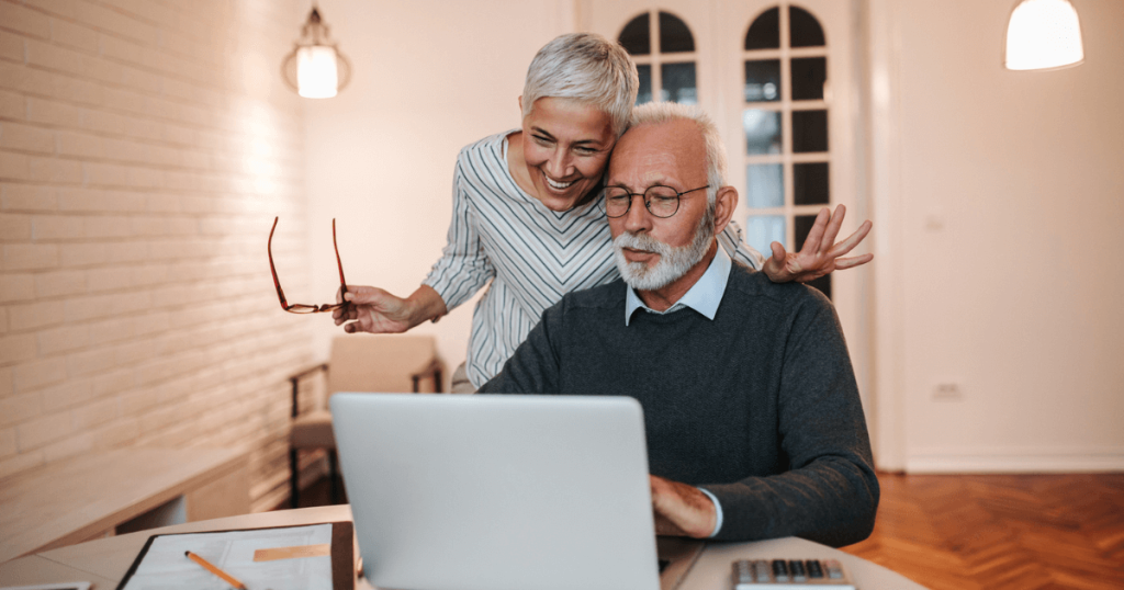 Older woman standing next to older man sitting down reviewing cost of assisted living on computer.