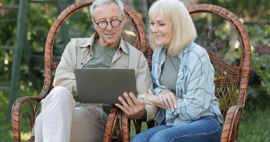 Elder man and woman sitting outside in chairs review the assisted living lifestyle on a laptop.